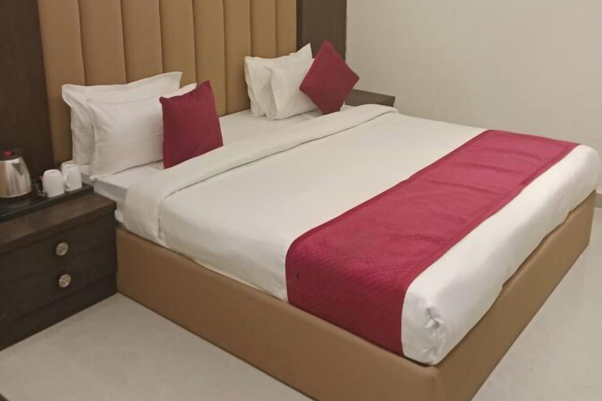 A neatly made bed with white and red bedding, a wooden nightstand with a kettle and cups, and a headboard with vertical slats.