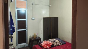 Una stanza piccola e luminosa con un letto, un armadio, una finestra dotata di zanzariera, una ventola a soffitto e un piccolo tavolo con oggetti personali.