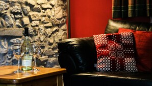 A stone wall, a red wall, a black leather sofa, and a wooden table with wine glasses and a bottle. - Yr Onnen, Aberaeron - Two Bedroom Cottage, Sleeps 4 (Llanarth)