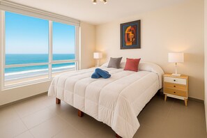 A hotel room with a large window offering a view of the beach and ocean, a bed with white bedding and two pillows, a bedside table with a lamp, and a framed artwork on the wall.