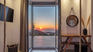 Ein Schlafzimmer mit einem großen Bett, einem Schreibtisch, einem Stuhl, einem hängenden Spiegel und Blick auf den Sonnenuntergang durch eine offene Tür.