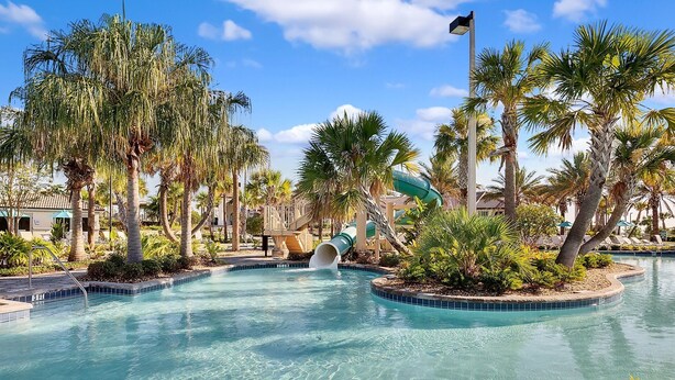A swimming pool with a water slide, surrounded by palm trees and lounge chairs.