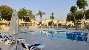 Ein Schwimmbad mit Liegestühlen und Sonnenschirmen, umgeben von Palmen und Gebäuden.
