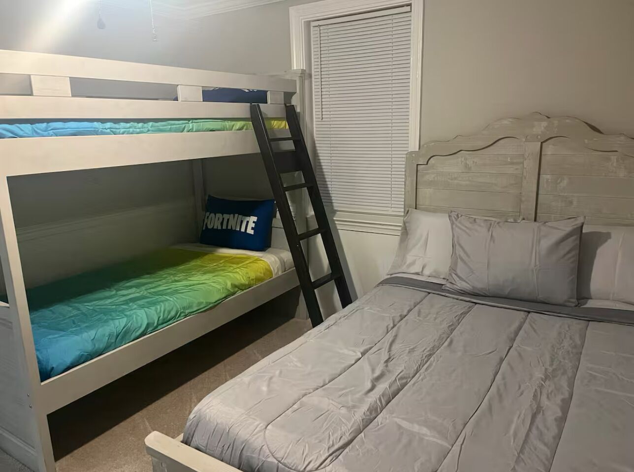 Ein Schlafraum mit Etagenbetten, einer Leiter, einem Fenster mit Jalousien und einem Bett mit Kissen.