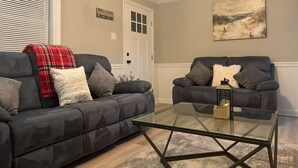 A living room with two dark grey sofas, a glass coffee table, and a beige wall with a landscape painting.