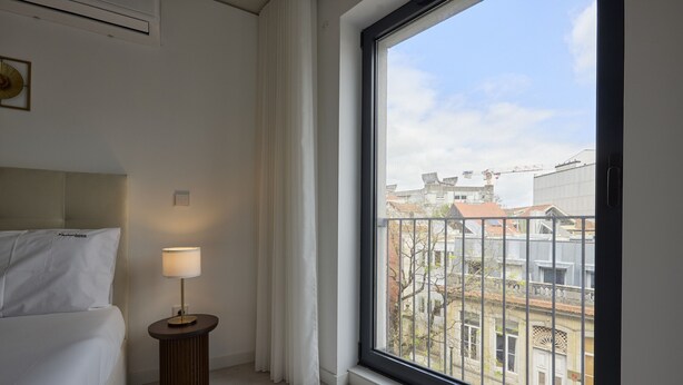 A hotel room with a bed, a bedside lamp, a window with a view of buildings, and a small table.