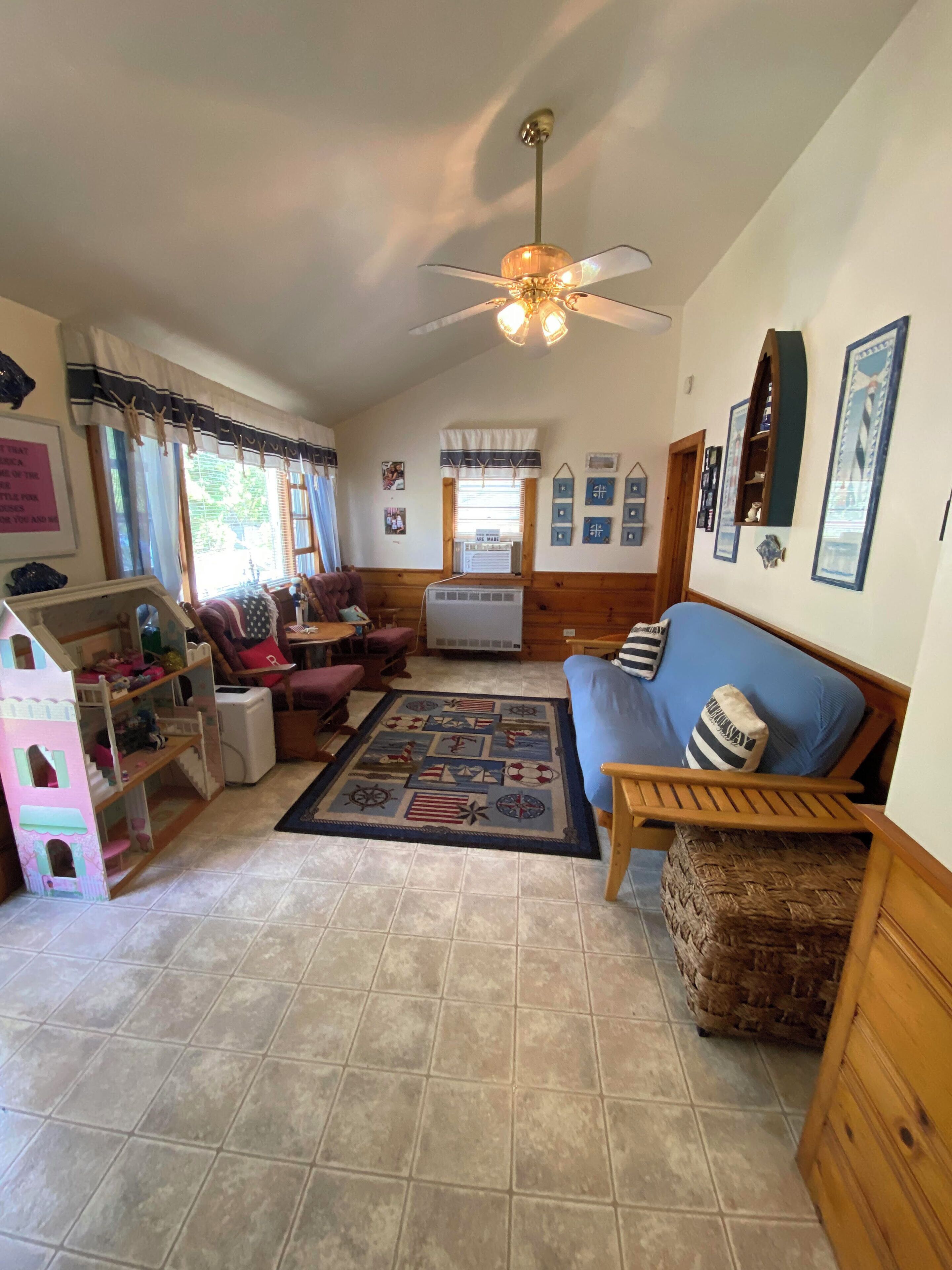 A cozy living room with a wooden floor, a blue sofa, a wooden coffee table, a ceiling fan, and a window with curtains.