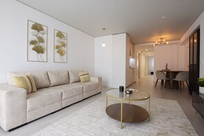 Een moderne woonkamer met een bank, een glazen salontafel en een eethoek op de achtergrond.