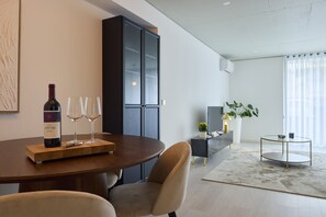 A modern dining area with a wooden table, wine bottle, and glasses. There is a dark cabinet, a plant, and a small round table with a lamp.