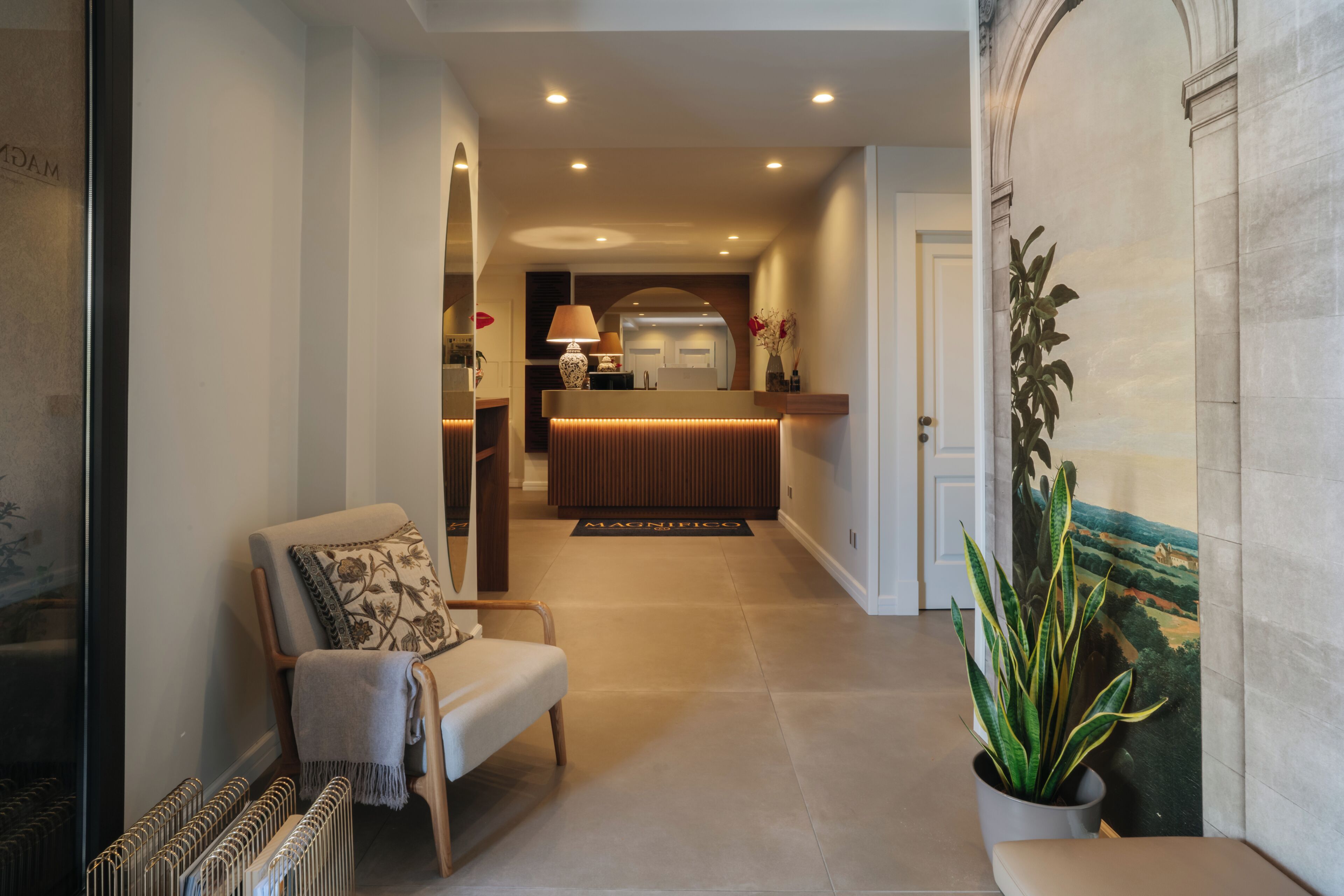 A hallway with a reception desk, a chair with a cushion, a potted plant, and a painting on the wall.