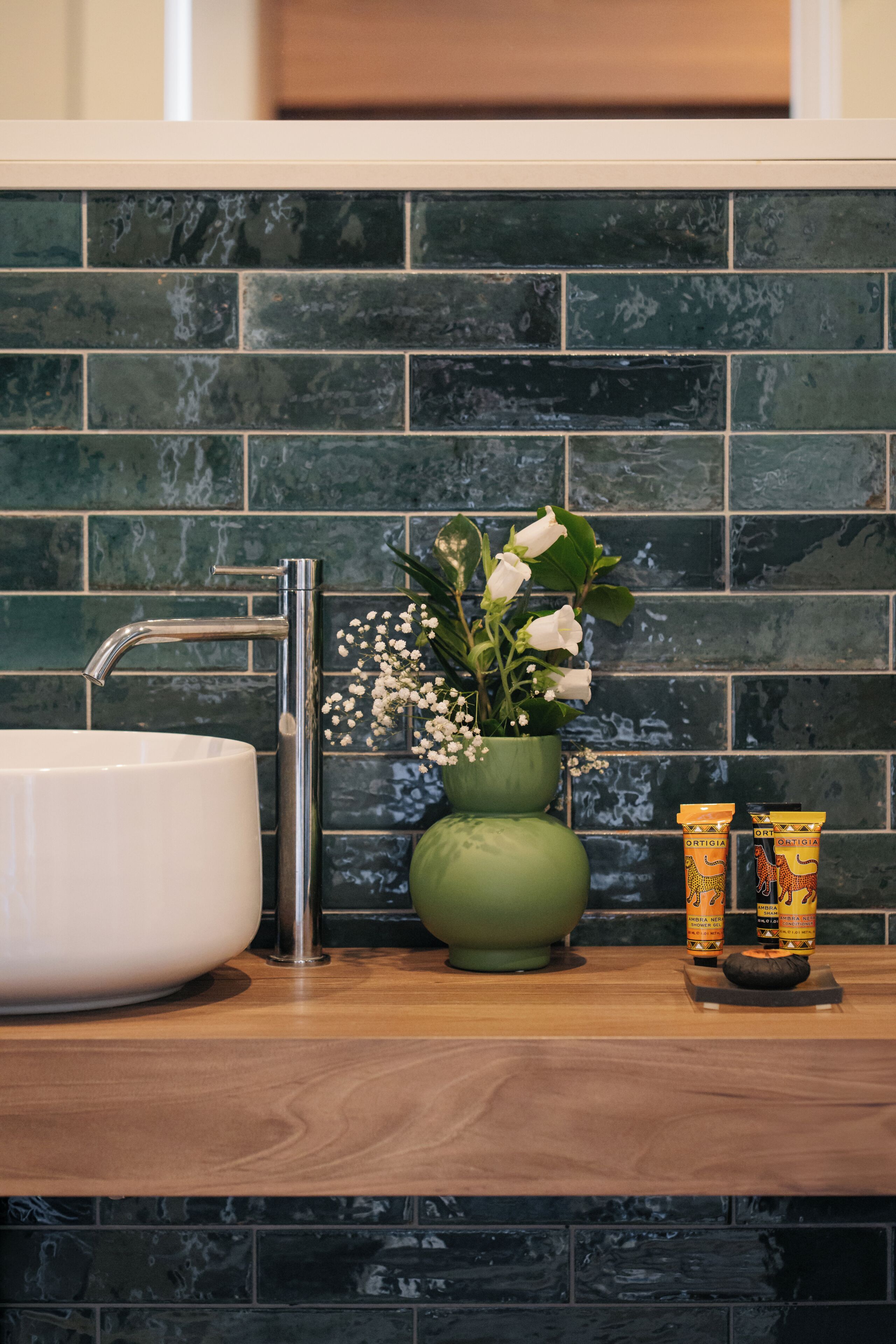 Un bagno con un lavandino bianco, un vaso verde con fiori e due contenitori per articoli da toilette sul ripiano.