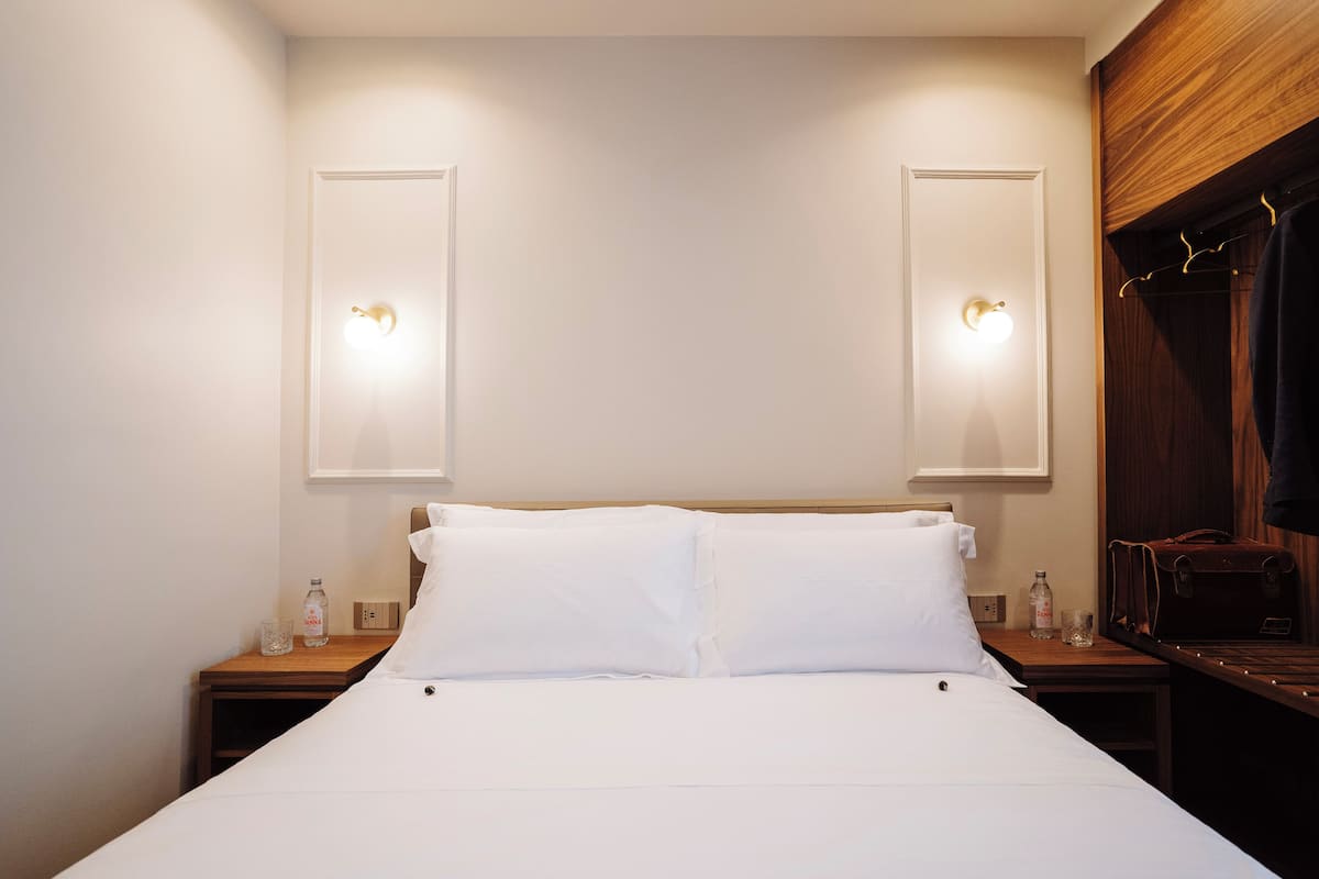 A neatly made bed with white linens, two bedside tables with lamps, and a wooden wardrobe with a hanging rack.