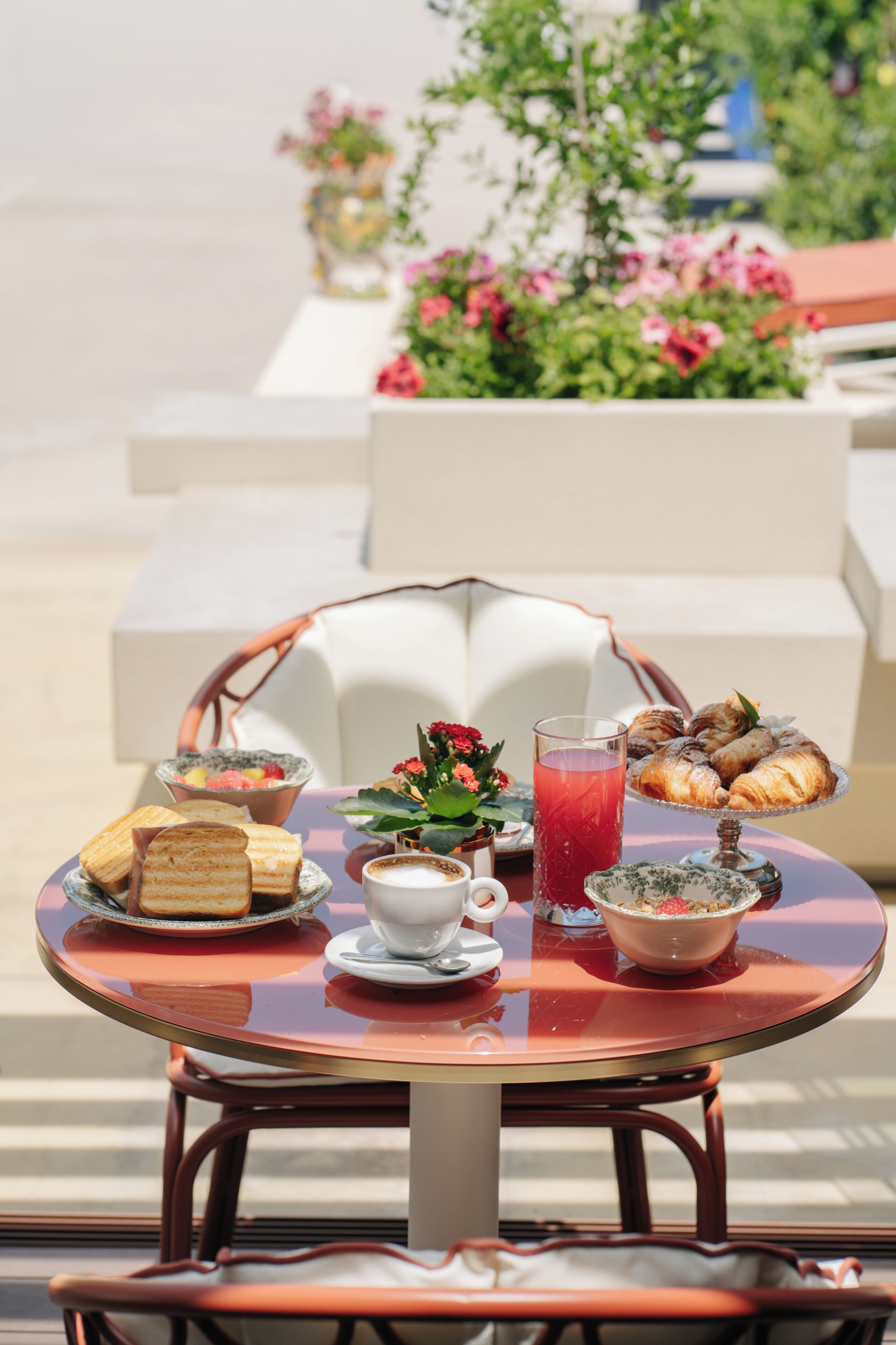 A round table with a variety of food items, a glass of red drink, and a white cup, set outdoors with a chair and a plant in the background.