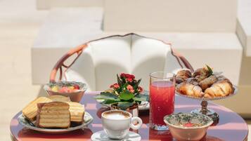 Una mesa redonda con diversos alimentos, un vaso de bebida roja y una taza blanca, ubicada al aire libre con una silla y una planta de fondo.
