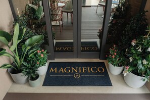 Entrada de un hotel con felpudo que dice 'MAGNIFICO', plantas en macetas y una vista del área de asientos interior.