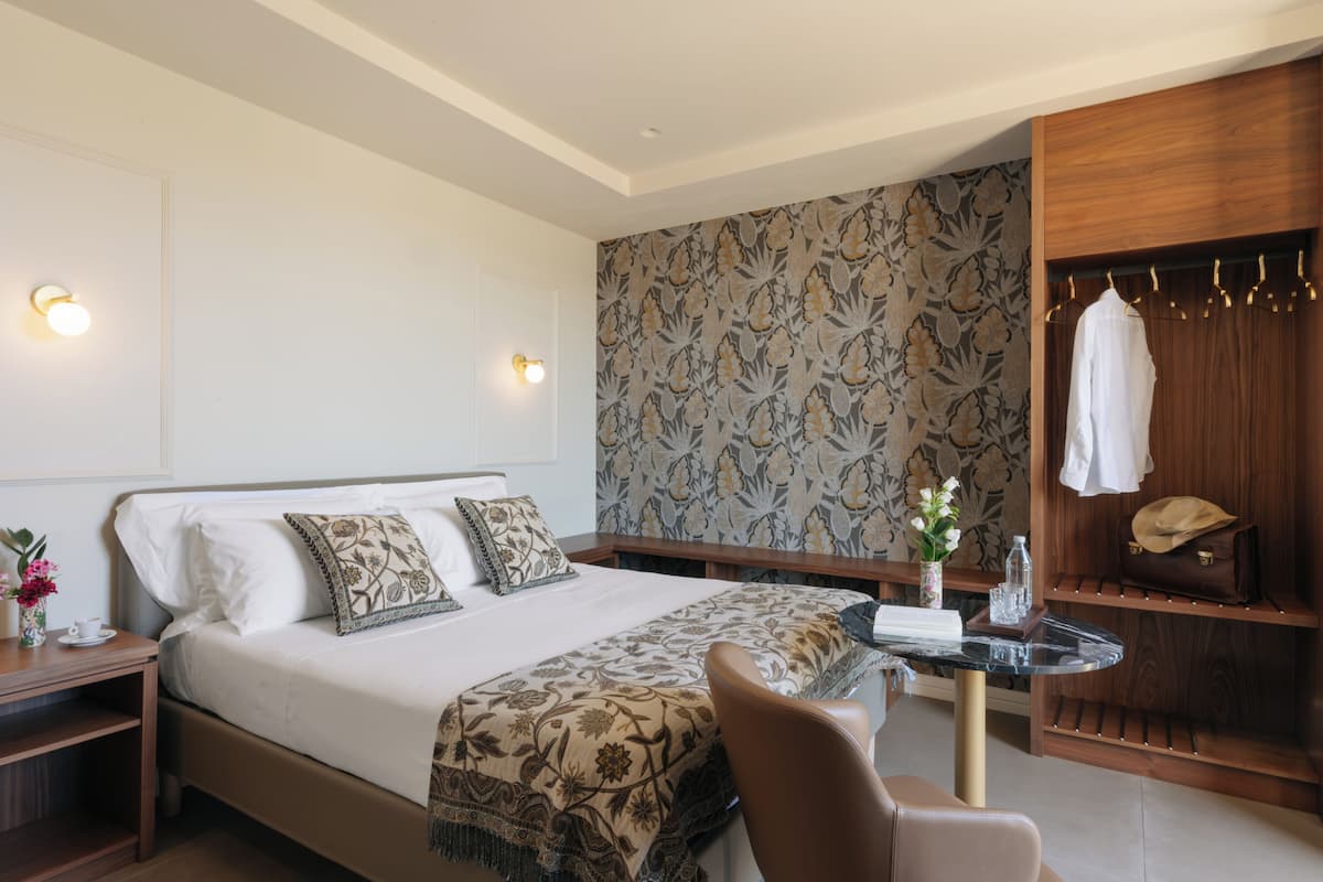A hotel room with a bed, bedside tables, a seating area, a vanity with a mirror, and a wardrobe with hanging clothes.