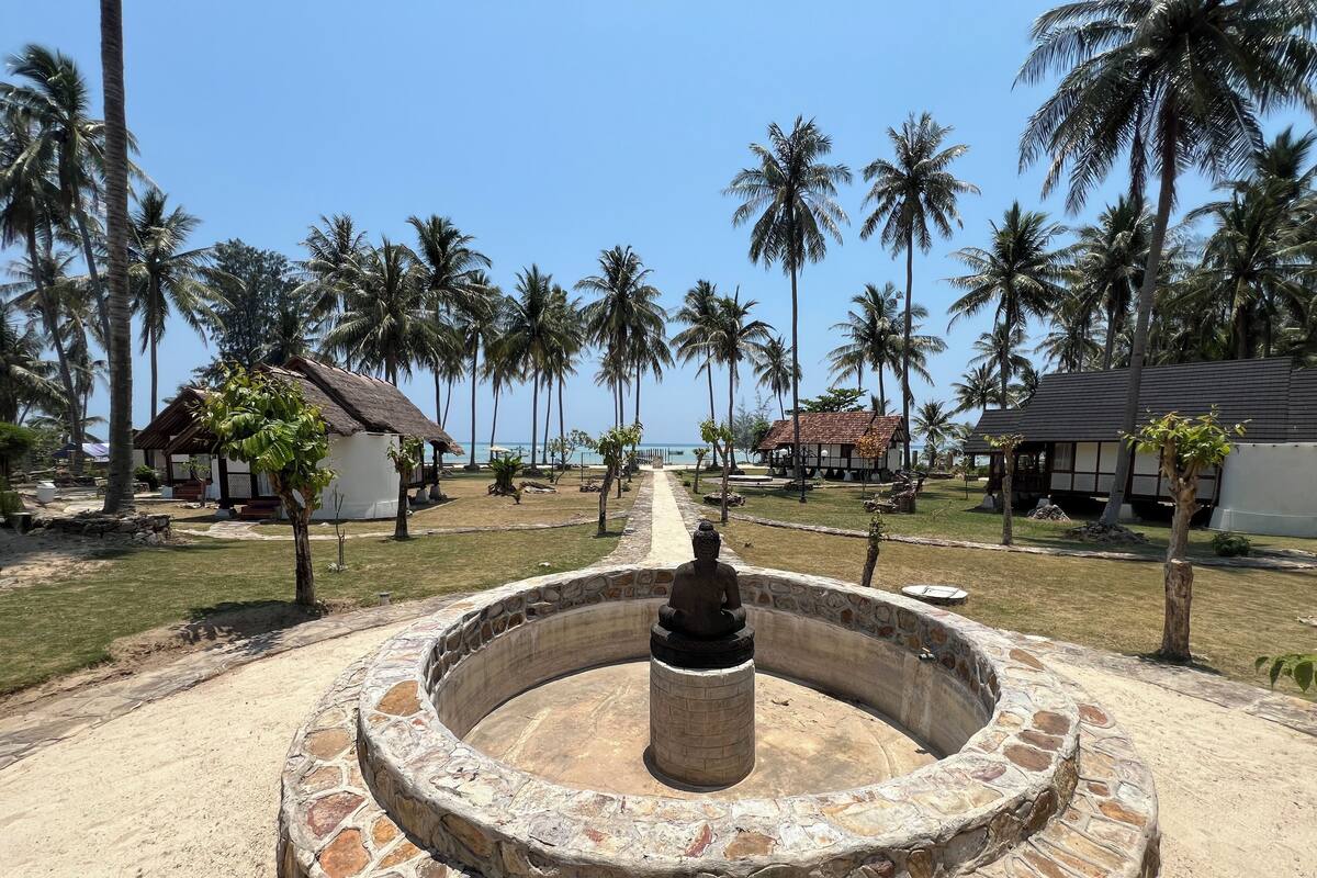 Una fuente de piedra con una estatua de Buda en el centro, rodeada por un muro de piedra, con palmeras y casas de playa al fondo.