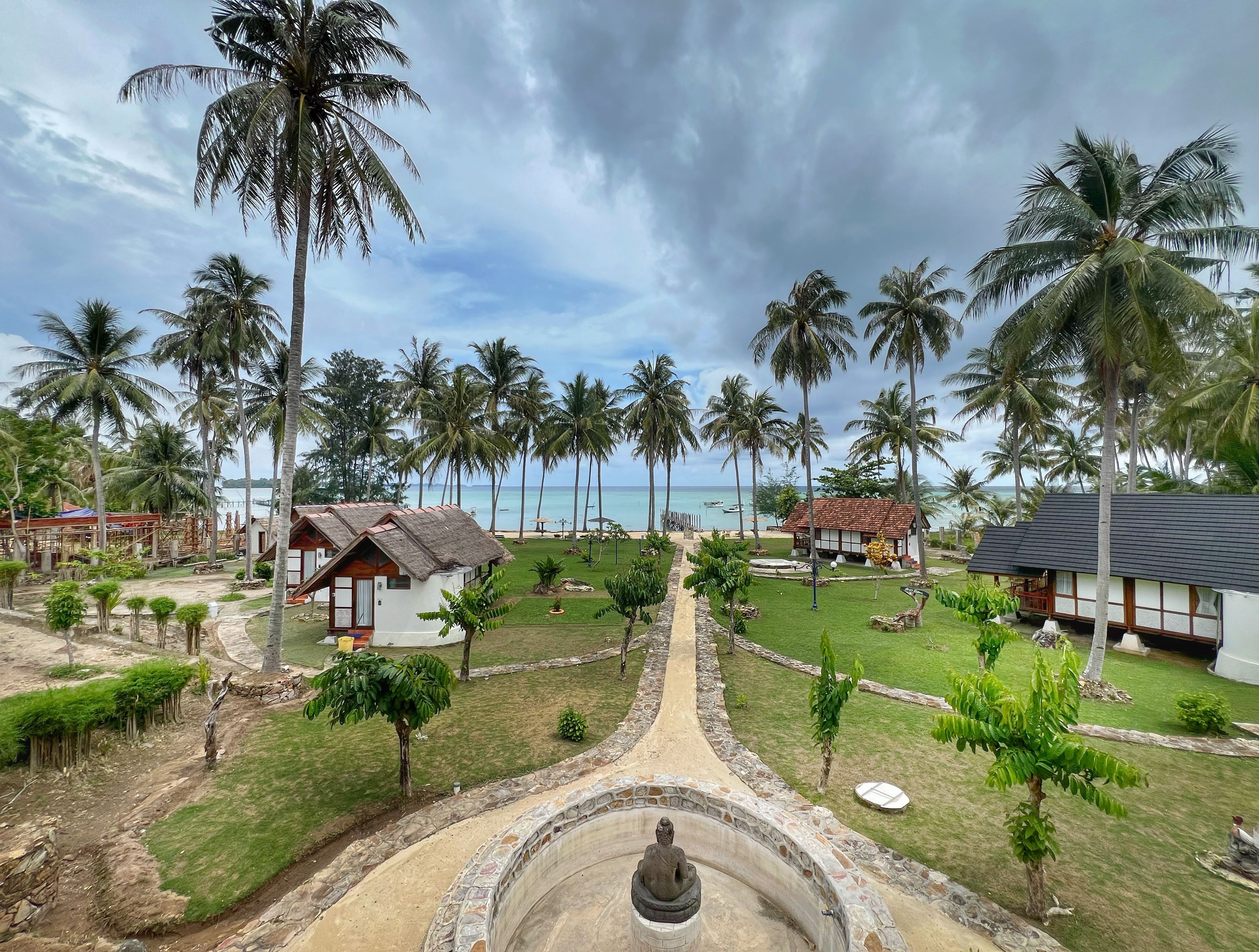 A resort with a central pathway leading to a statue, surrounded by palm trees and small buildings.