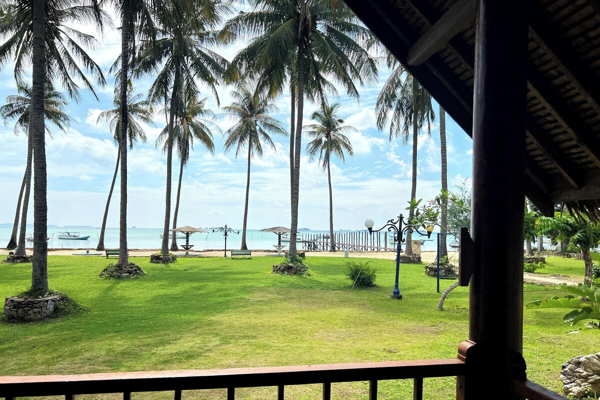A view from a balcony overlooking a beach with palm trees, a bench, and a boat in the distance.