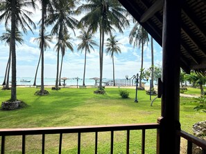 Vista desde un balcón sobre una playa con palmeras, un banco y un barco a lo lejos.