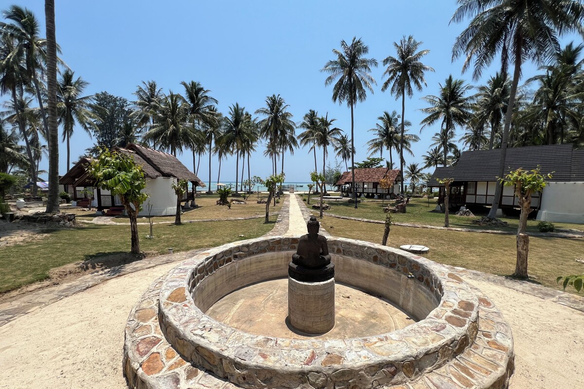 Una fuente de piedra con una estatua de Buda en el centro, rodeada por un muro de piedra, con palmeras y casas de playa al fondo.
