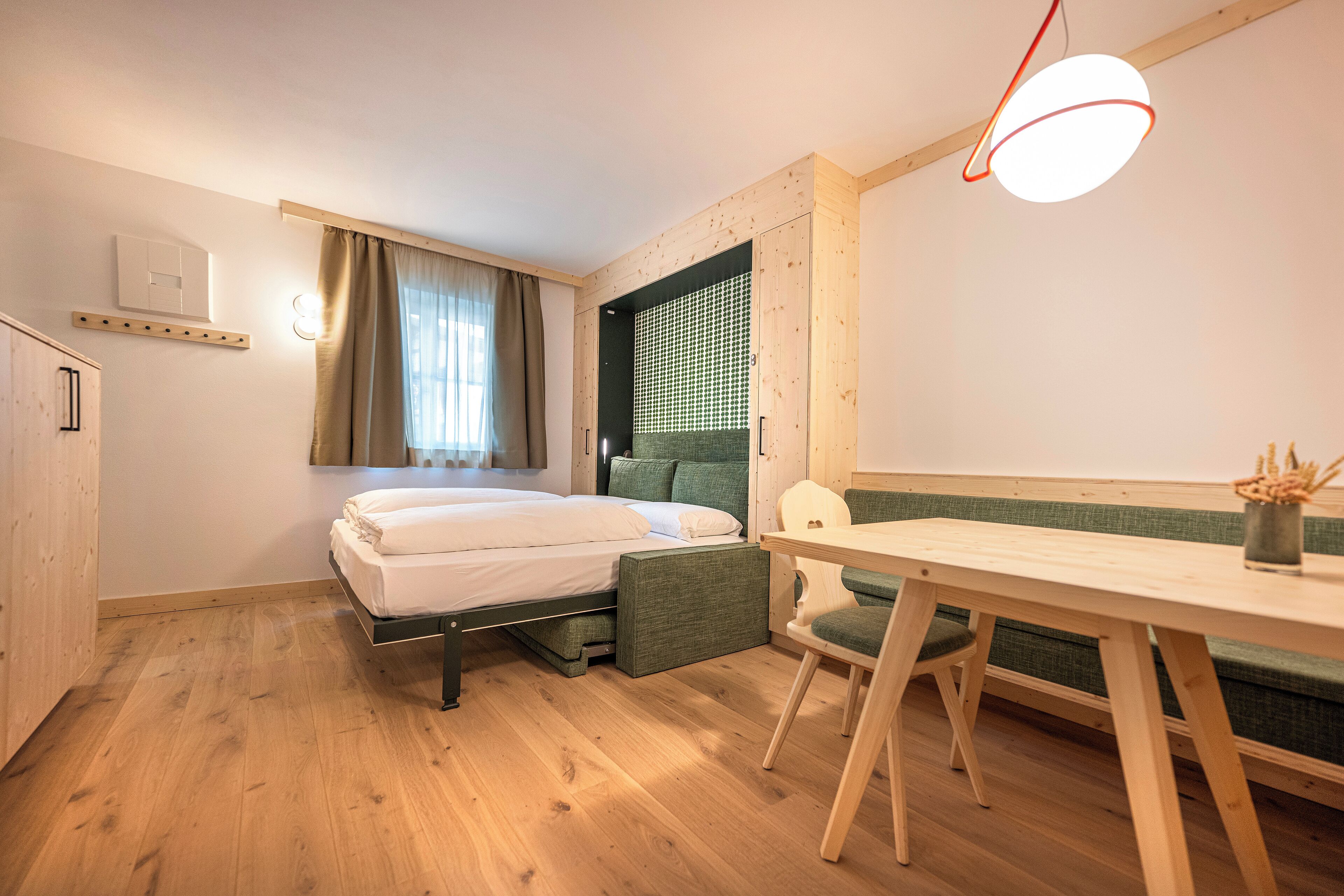 Een kleine kamer met een bed, een bureau en een stoel. Er is een raam met gordijnen, een wandplank en een plafondlamp.