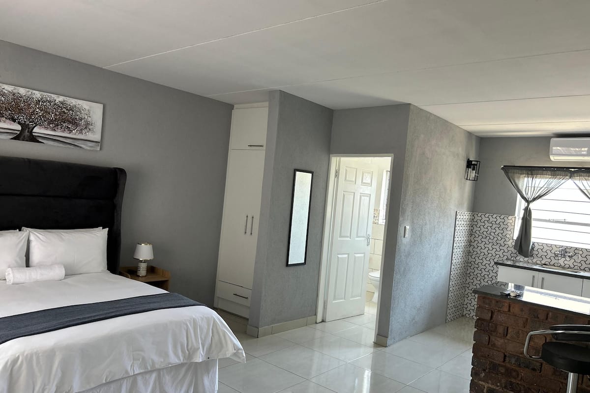 Une chambre avec un grand lit, une armoire, une salle de bain visible par une porte ouverte et un coin cuisine avec un mur de fond en briques.
