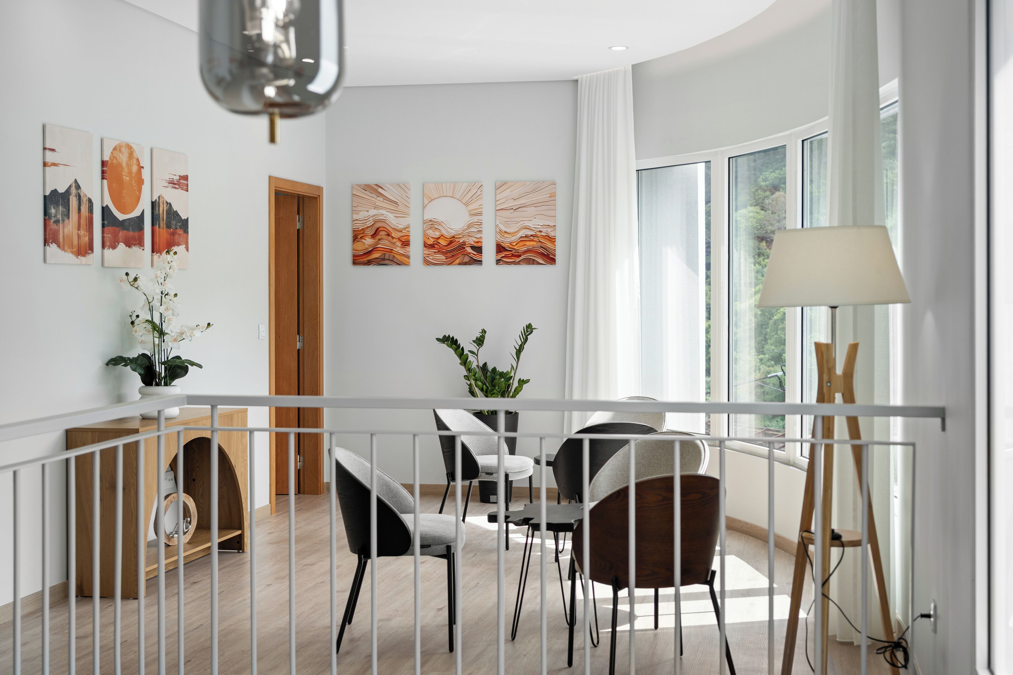 A modern dining area with a white railing, a wooden table, and chairs. There are three paintings on the wall, a potted plant, and a lamp.