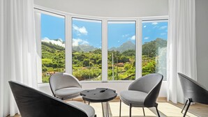 Una habitación con ventanal panorámico que da a un paisaje exuberante. Hay dos sillones grises y una mesita redonda.