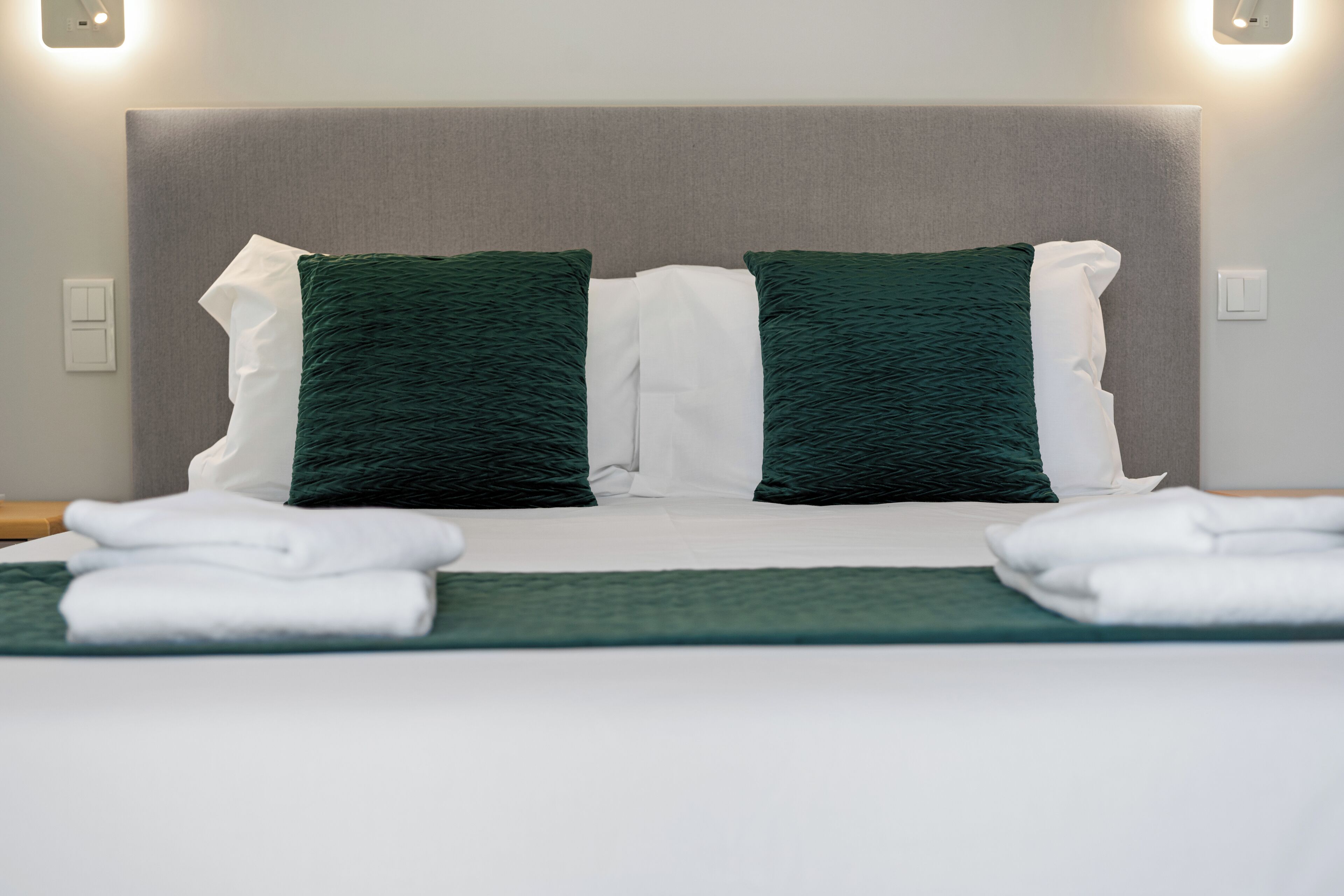 Una cama bien hecha con ropa de cama blanca y verde, un cabecero gris y dos lámparas fijadas a la pared.