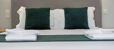 Una cama bien hecha con ropa de cama blanca y verde, un cabecero gris y dos lámparas fijadas a la pared.