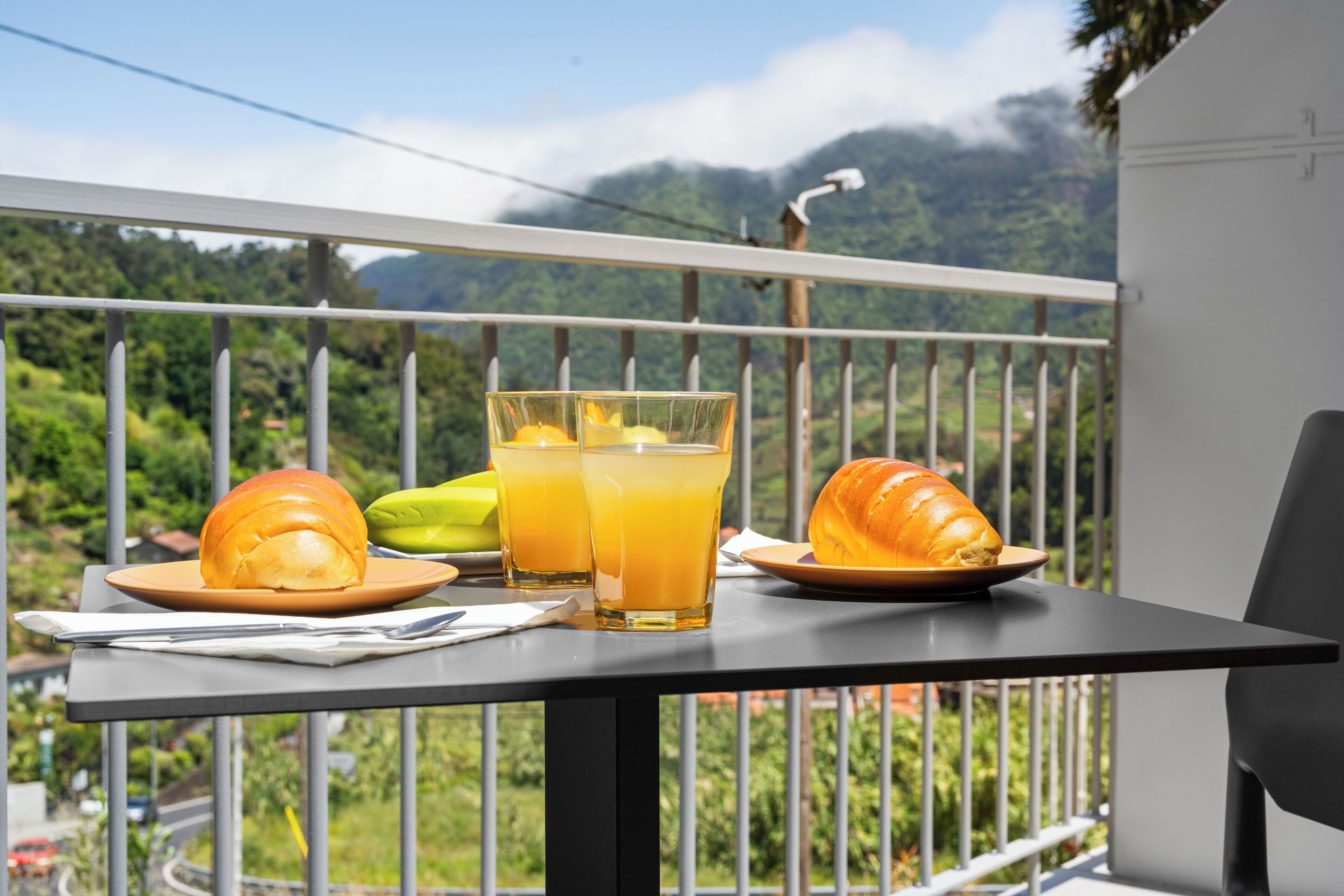 A balcony with a table set for breakfast, including a croissant, a sandwich, a banana, and two glasses of orange juice.
