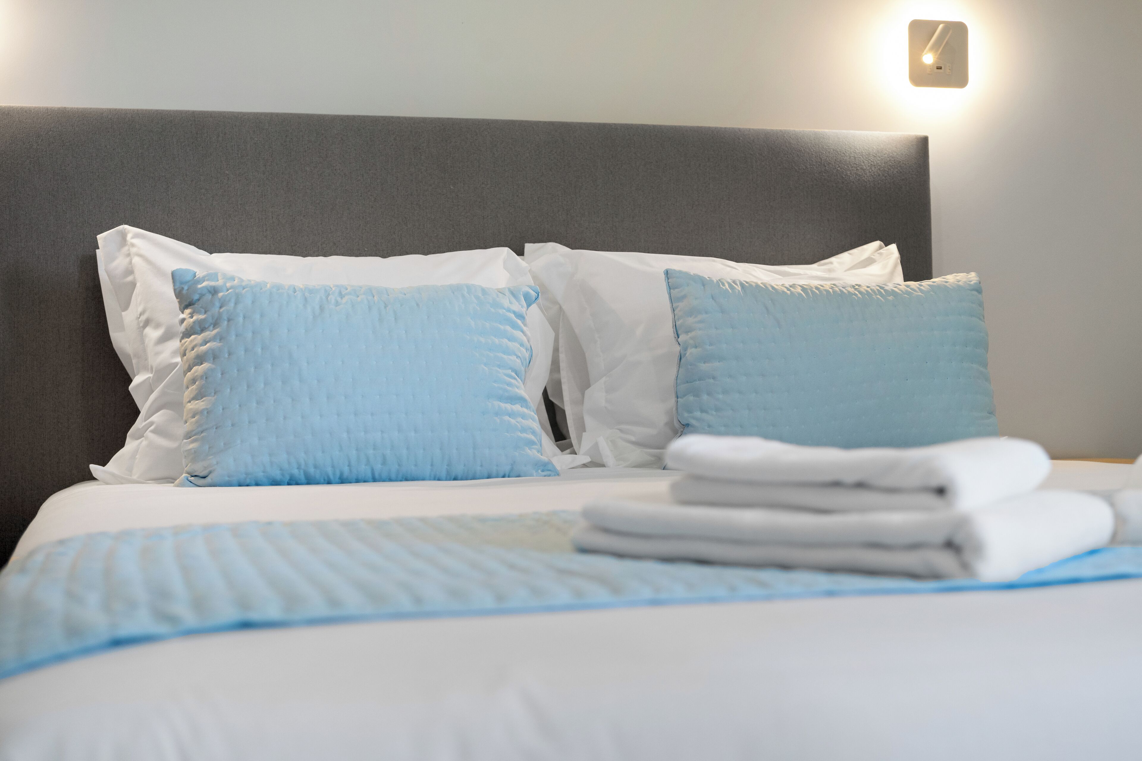 Ein ordentlich bezogenes Bett mit weißer und blauer Bettwäsche, ein gefaltetes weißes Handtuch und eine wandmontierte Leuchte.