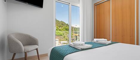 A hotel room with a bed, a chair, a TV, a balcony with a view of greenery, and a wooden wardrobe.