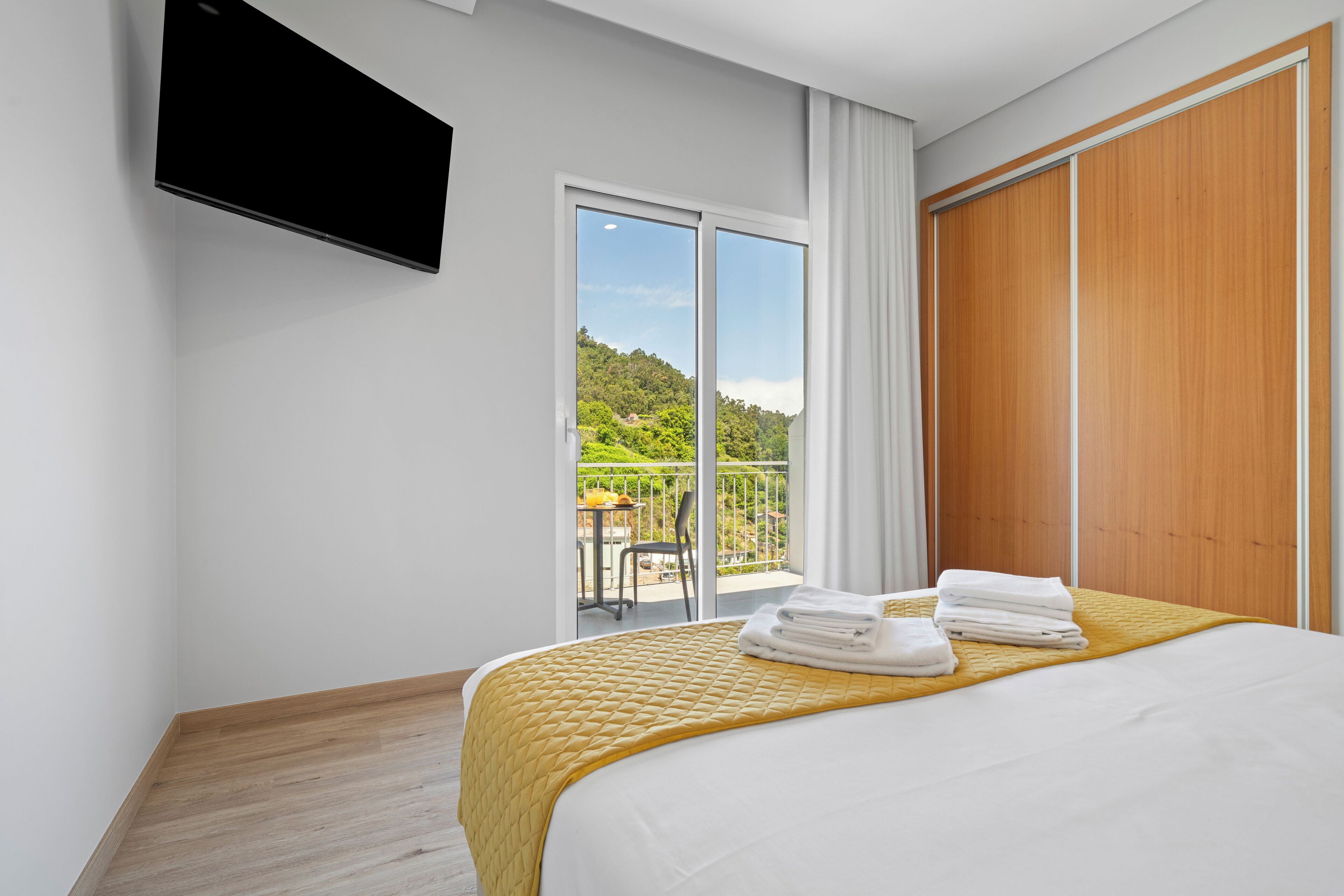 A hotel room with a bed, a TV mounted on the wall, and a balcony with a view of greenery.