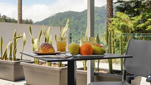 Una mesa con frutas y una bebida, rodeada de plantas en macetas y una silla, con vistas a la montaña al fondo.