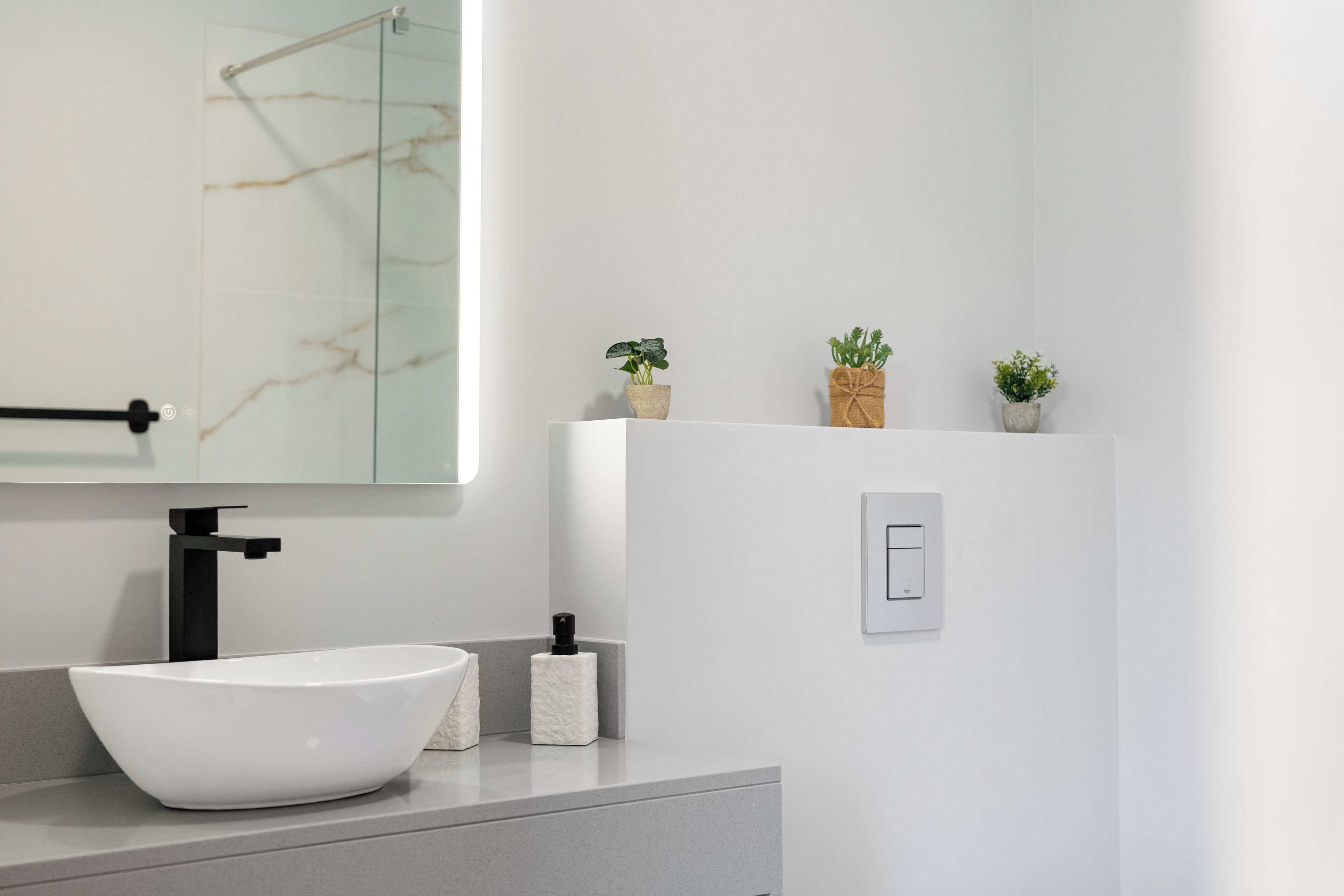 A modern bathroom with a white sink, a large mirror, and a marble backsplash. There are three small potted plants on a shelf above the sink.