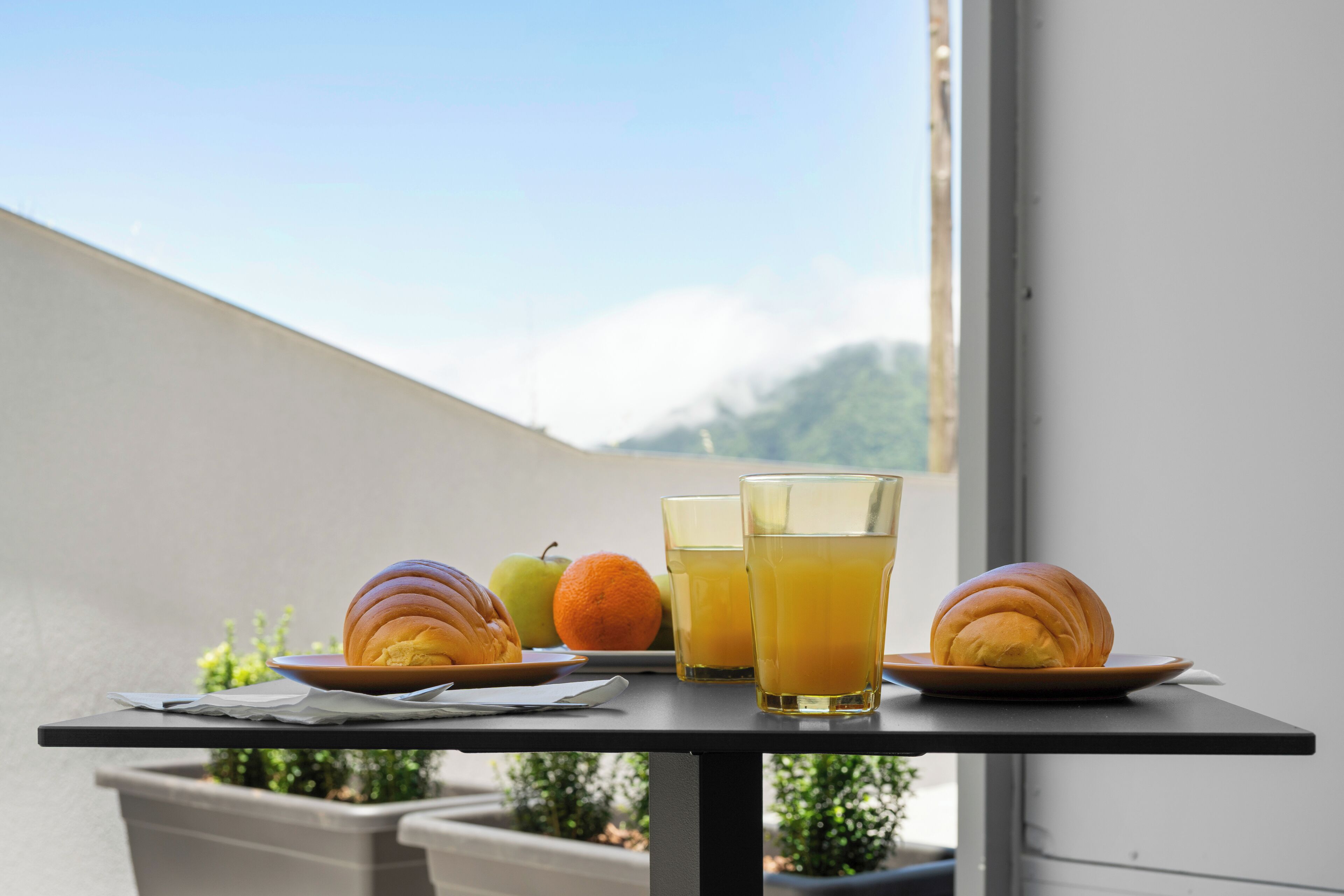 Een tafel met croissants, fruit en glazen sinaasappelsap op een balkon met uitzicht op bergen.