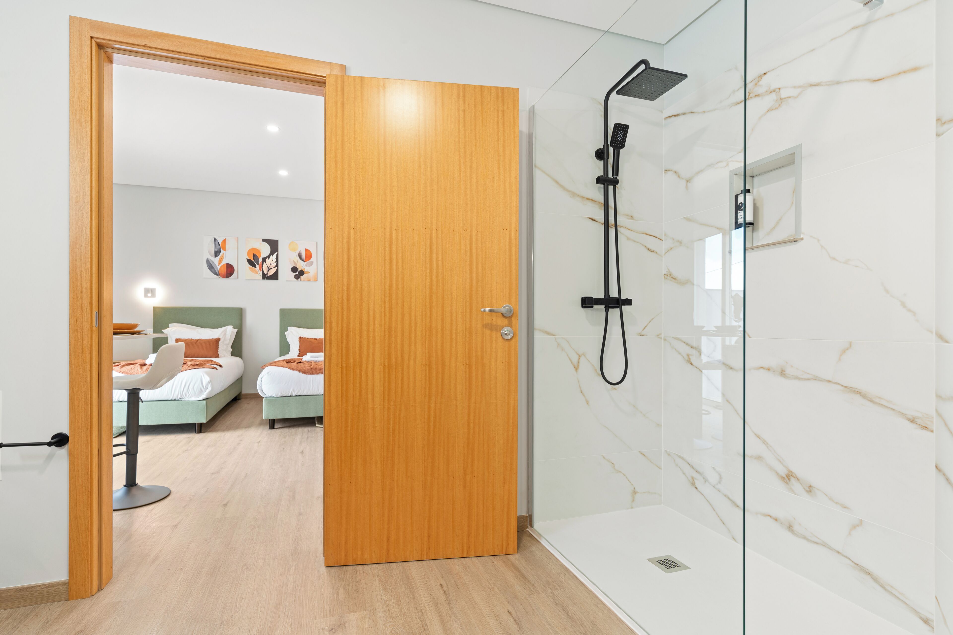 Habitació d'hotel moderna amb una porta de fusta, una dutxa amb plat de rodet i dos llits amb capçaleres i coixins a conjunt.