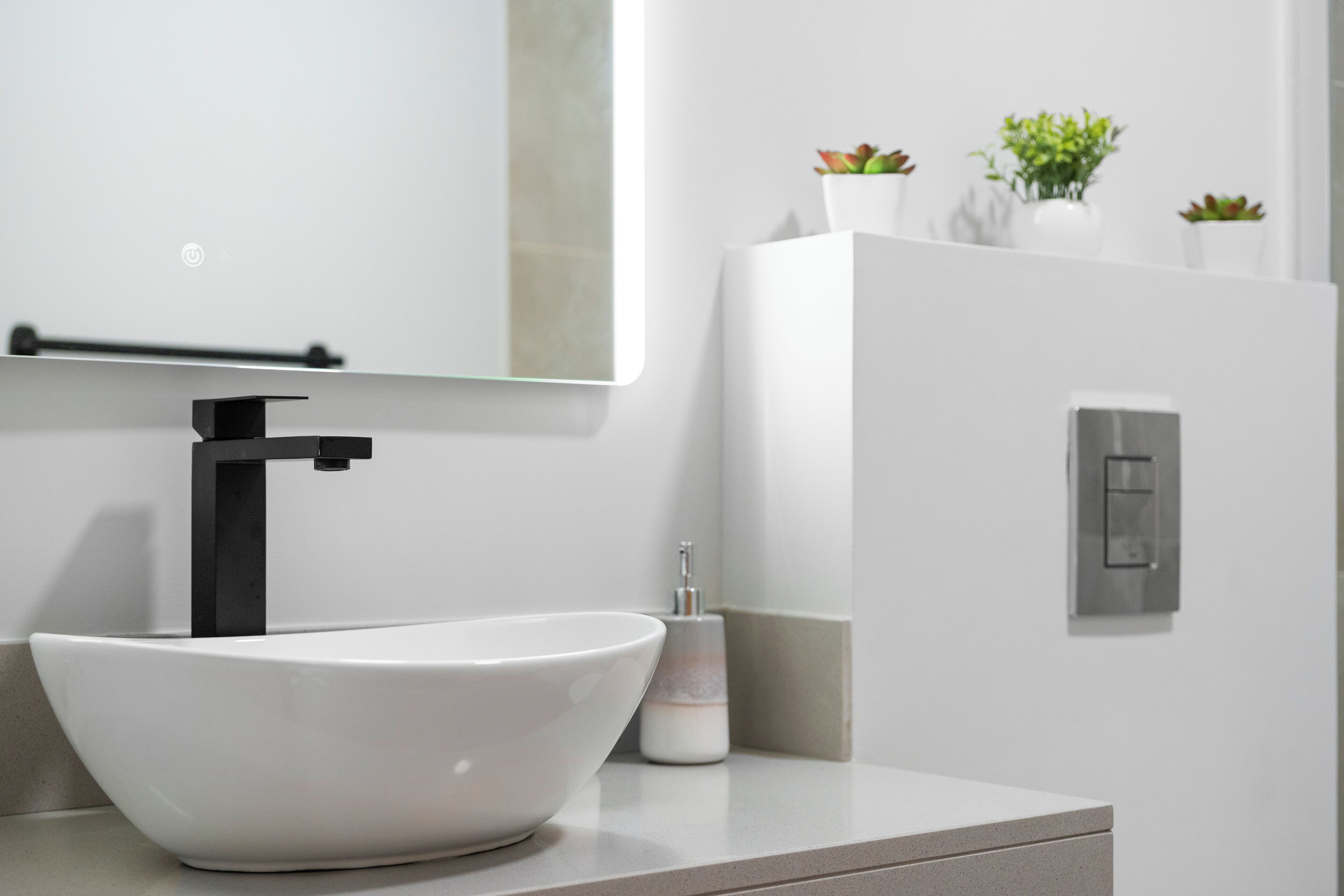 A modern bathroom with a white sink, black faucet, and a mirror above it. There are three potted plants on the wall and a light switch to the right.