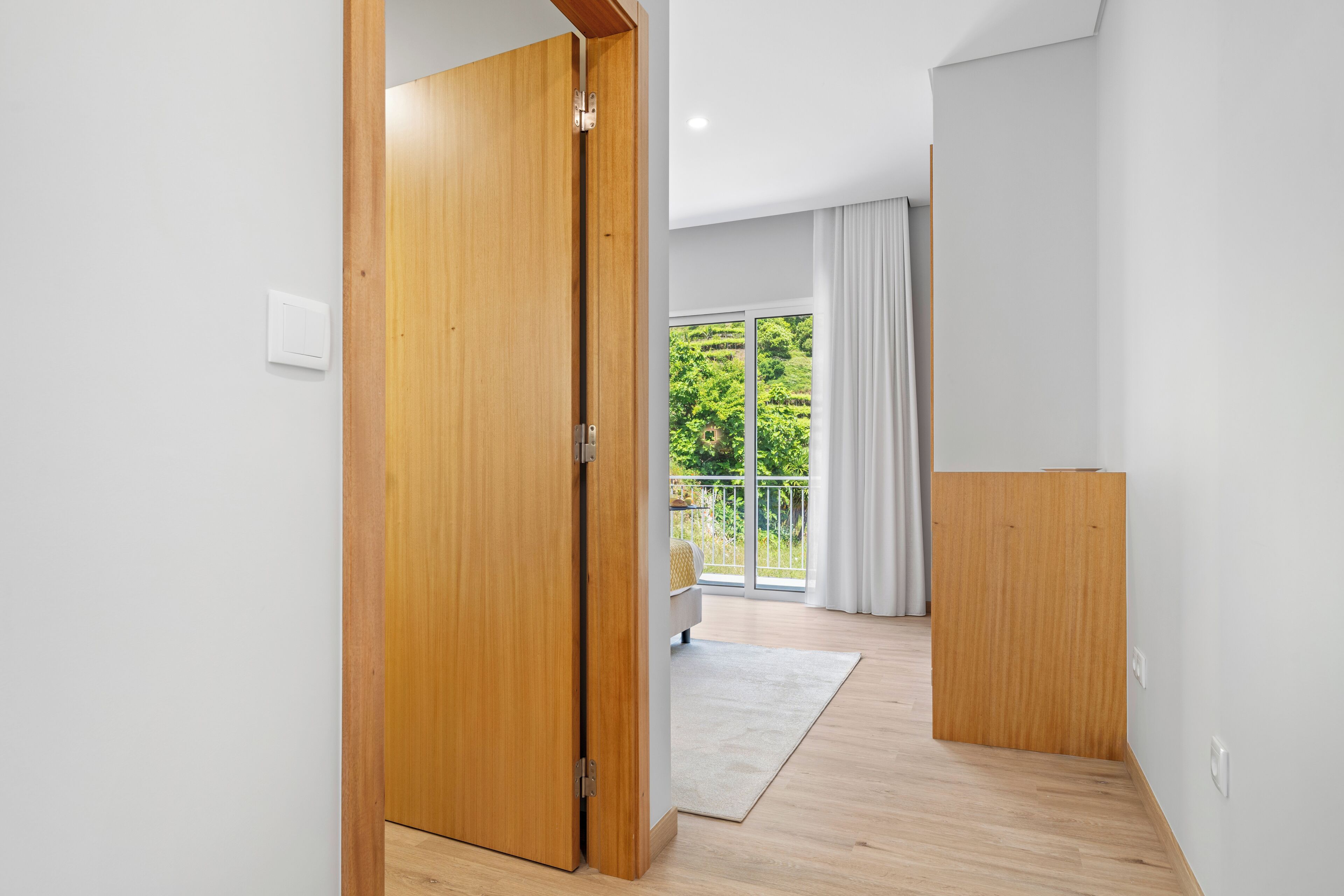 A modern interior with a wooden door, a view of a balcony with greenery, and a light-colored floor.