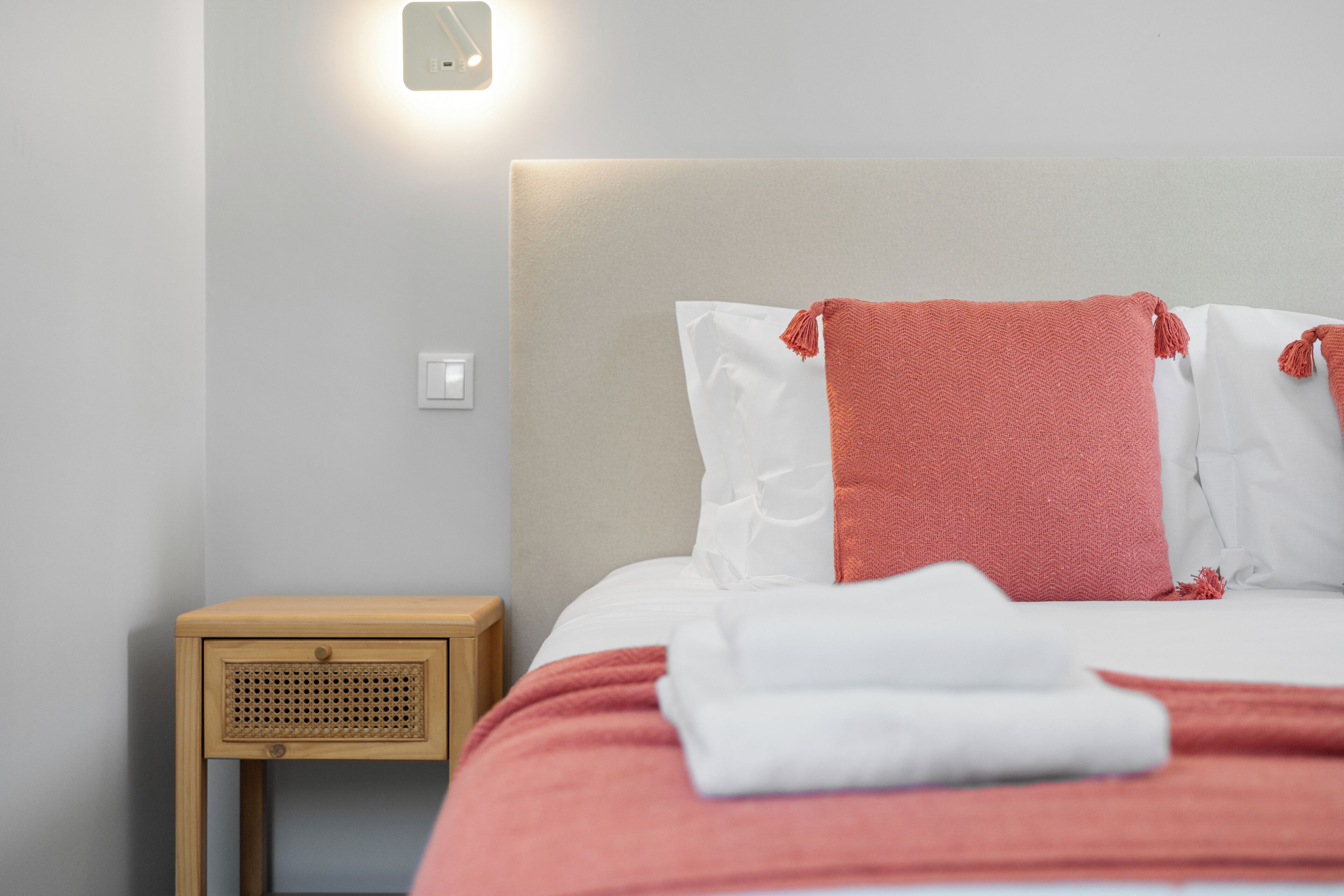 Una cama bien tendida con ropa de cama blanca y rosa, una mesita de noche de madera con una cesta tejida y una lámpara de pared.
