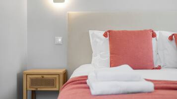 Una cama bien tendida con ropa de cama blanca y rosa, una mesita de noche de madera con una cesta tejida y una lámpara de pared.