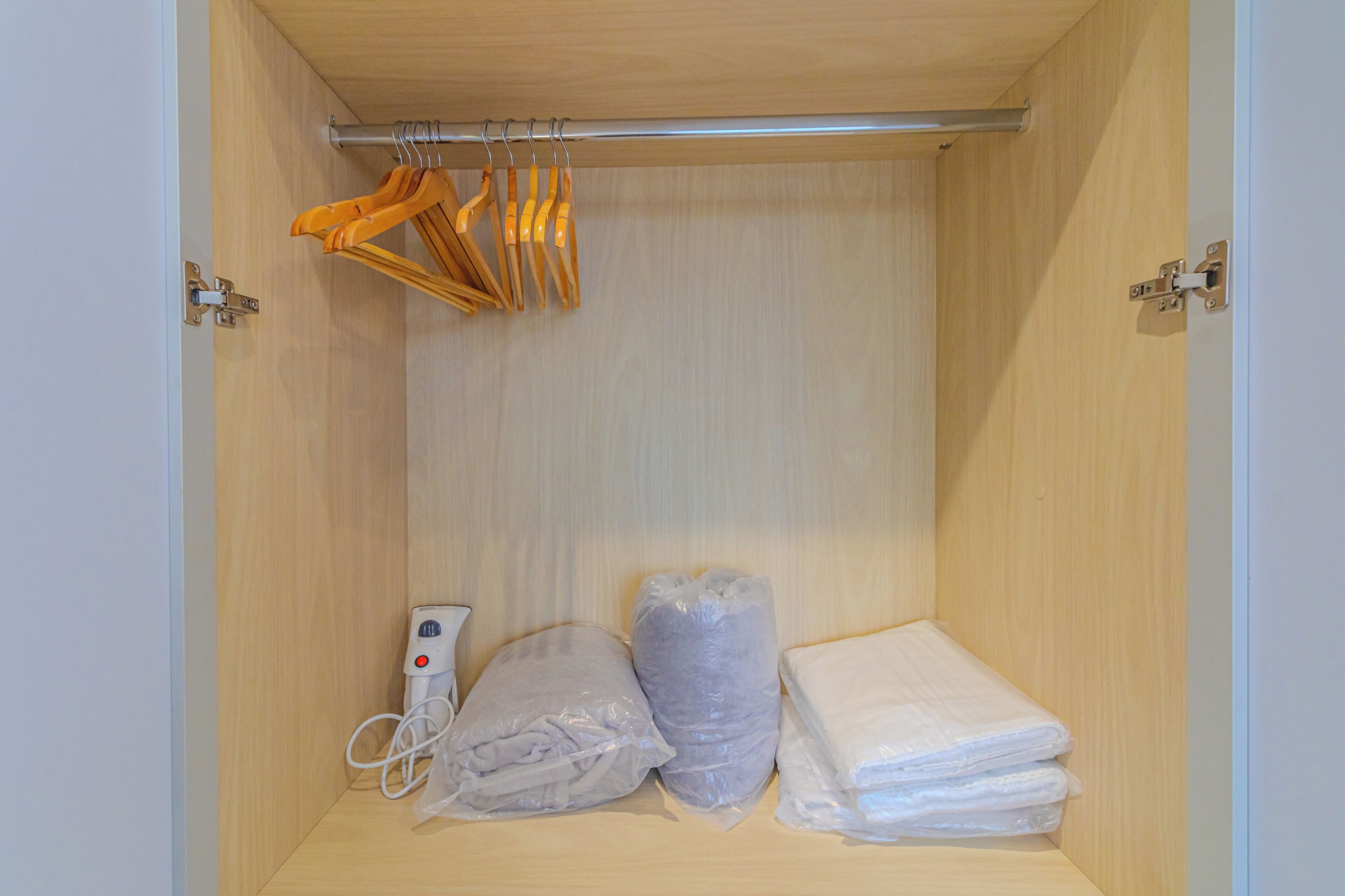 A small, wooden closet with a metal rod for hanging clothes, a white device, and two plastic-wrapped items.