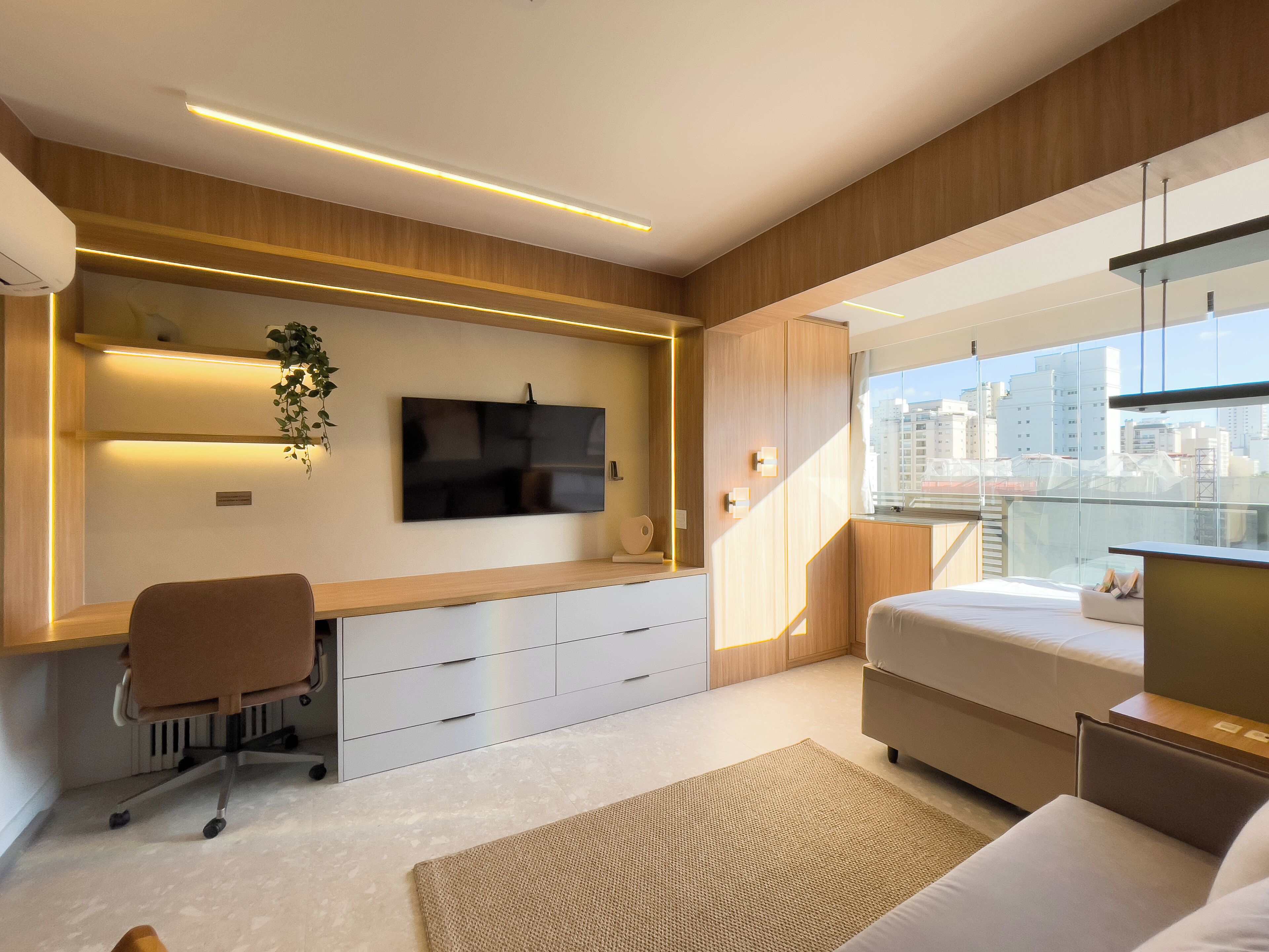 A modern bedroom with a large bed, a desk with a chair, a TV mounted on the wall, and a city view through large windows.