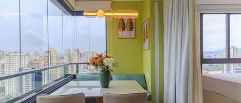 A balcony with a glass railing, a table with chairs, a vase with flowers, and a city view.