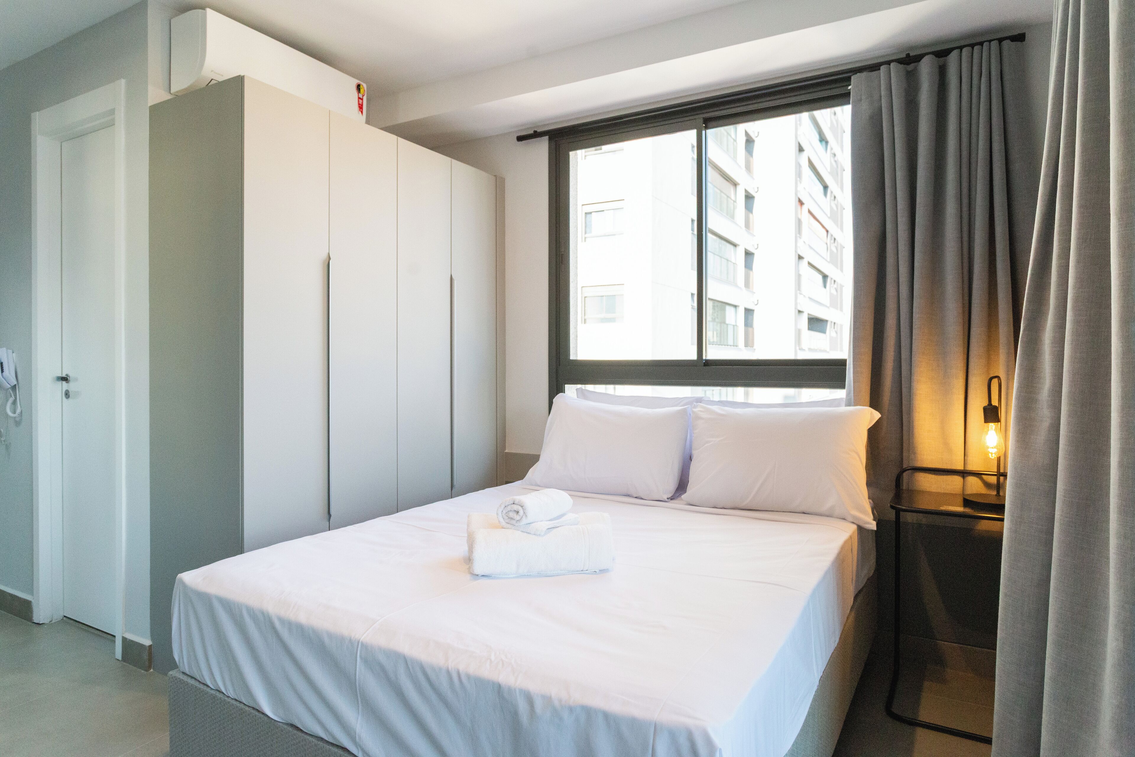 Una habitación de hotel con cama, armario, mesita de noche con lámpara y ventana con cortinas.