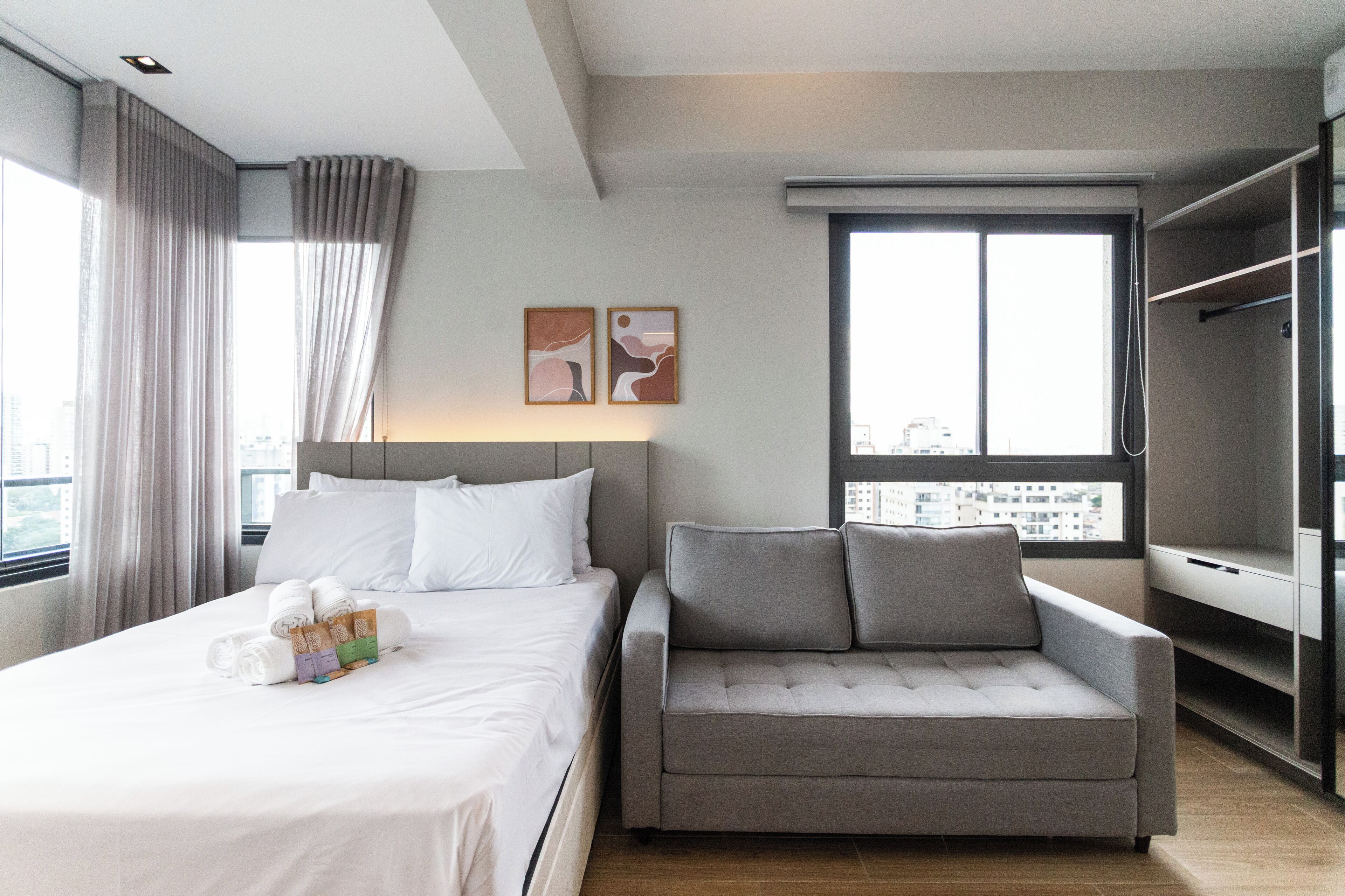 Quarto de hotel moderno com uma cama bem arrumada, um sofá cinzento e uma grande janela com vista para a cidade.
