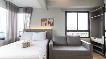 Quarto de hotel moderno com uma cama bem arrumada, um sofá cinzento e uma grande janela com vista para a cidade.