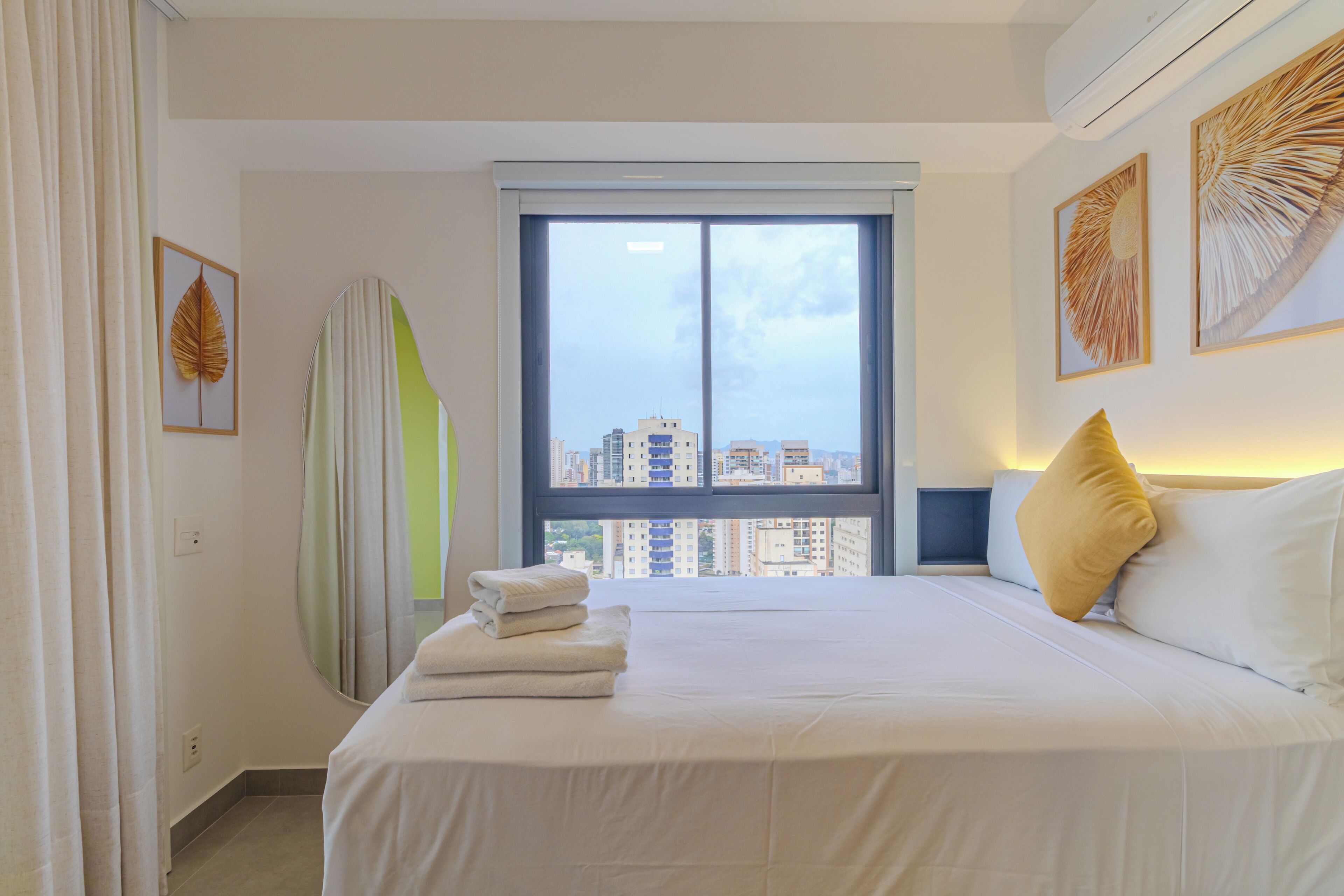 A hotel room with a bed, a window with a city view, and artwork on the walls.